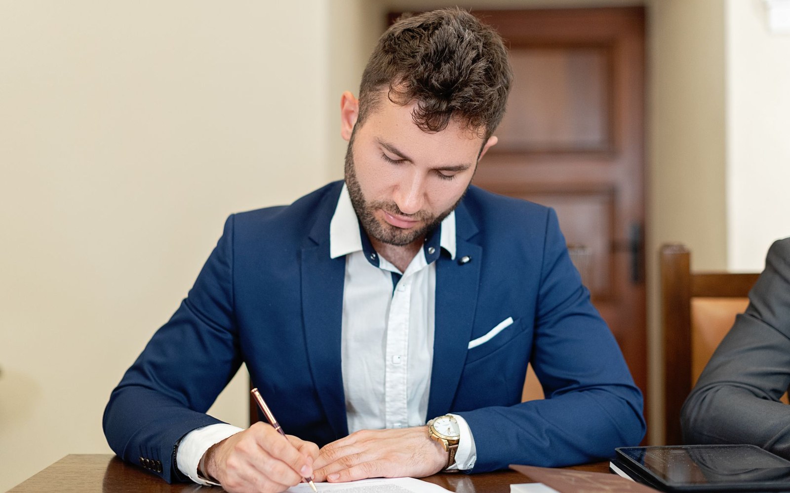 Man in a blue blazer signing a document at a wooden desk