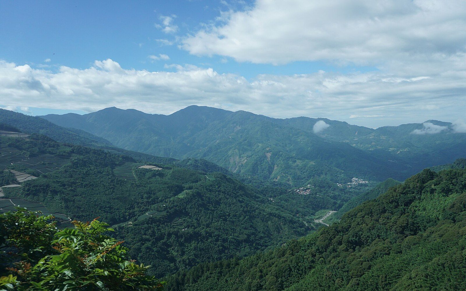 Forested mountain ridges and a valley village stretch beneath a partly cloudy sky