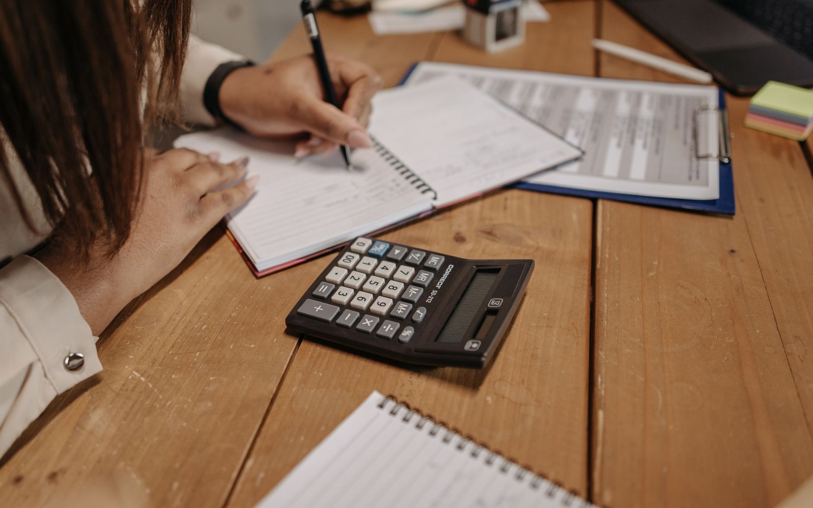A person writes in a notebook beside a calculator and clipboard on a wooden desk