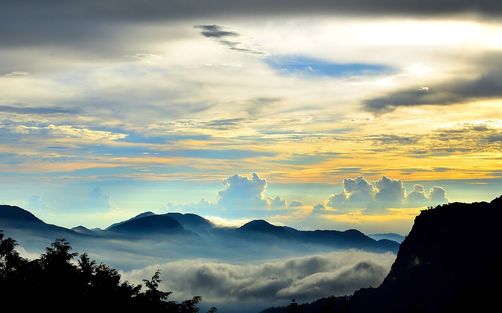 Layered mountain ridges rise above a cloud inversion beneath a colorful sunset sky